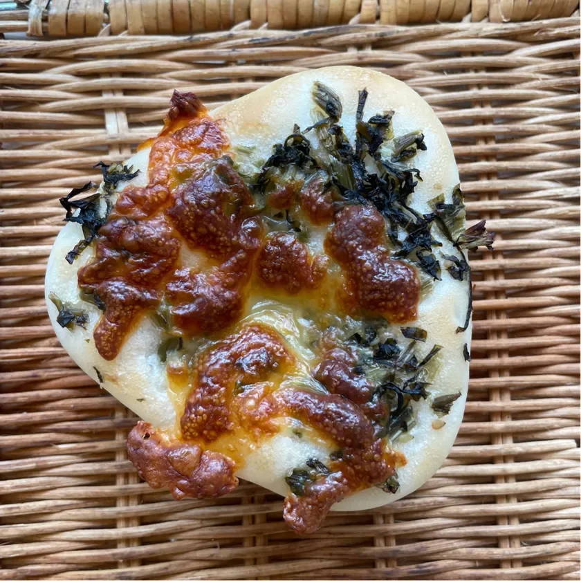 籐のマットの上に置かれた、焼き色のついたチーズと緑の葉物野菜がトッピングされた小さなパンです。香ばしく焼けたチーズと具材が食欲をそそります。