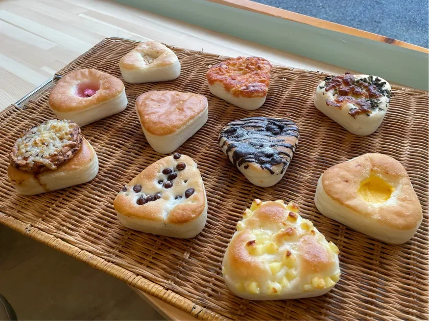 様々な種類のパンが籐のバスケットに並べられています。甘いものからしょっぱいものまで、三角形や丸みを帯びた形のパンがあり、それぞれ異なるトッピングが施されています。