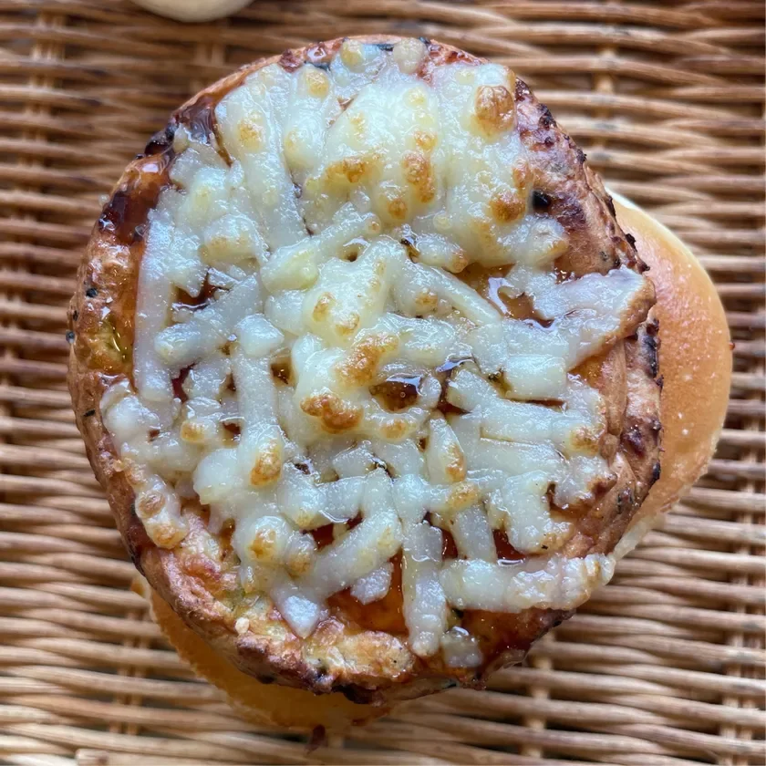 籐のバスケットに置かれた、香ばしく焼き上げられた丸い惣菜パンのアップです。表面にはとろけるチーズとソースがたっぷりとかかっており、食欲をそそる一品です。