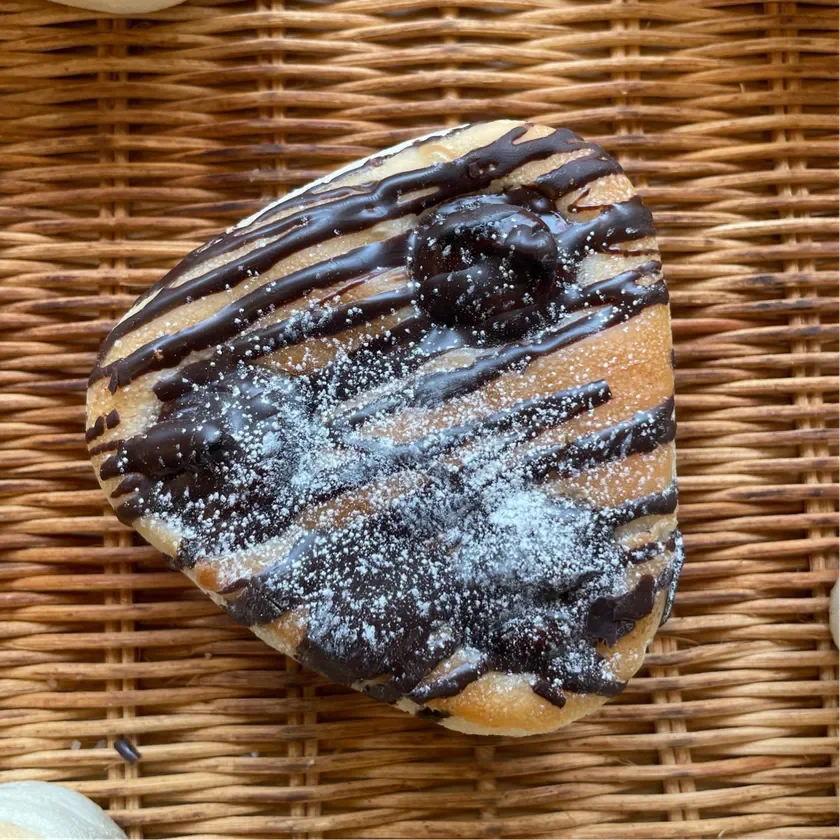 籐製のバスケットに置かれた、チョコレートソースと粉砂糖で飾られた菓子パンのクローズアップ画像です。甘く焼かれた生地に、とろけるようなチョコレートの層が食欲をそそります。