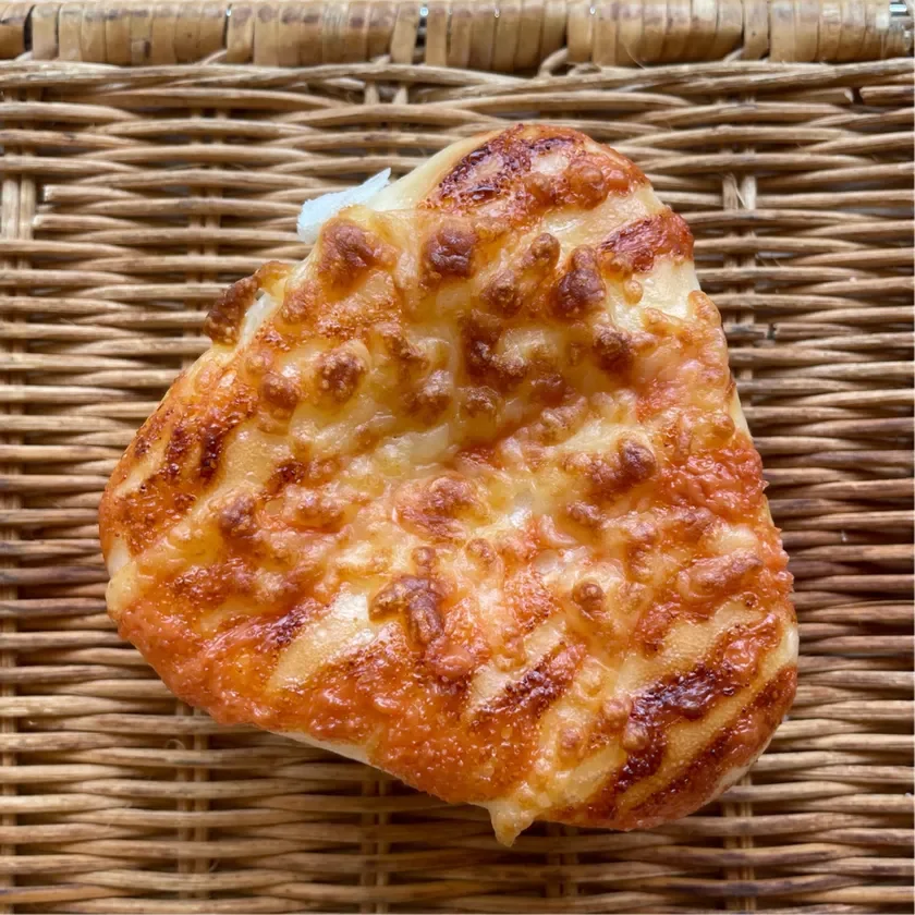 ラタンの編み込みの上に置かれた、香ばしく焼き上げられたチーズパンのクローズアップ。とろけたチーズがたっぷり乗っており、一部はカリカリに焼けていて食欲をそそる。
