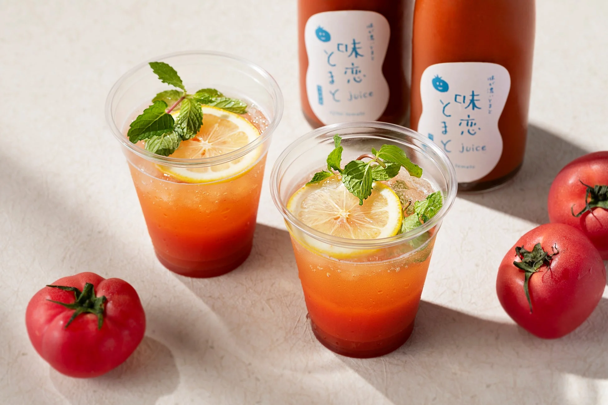 トマトジュースを使った爽やかなドリンクが2杯、フレッシュなトマトとボトルジュースと共に並べられています。レモンとミントが添えられ、夏らしい清涼感を演出しています。