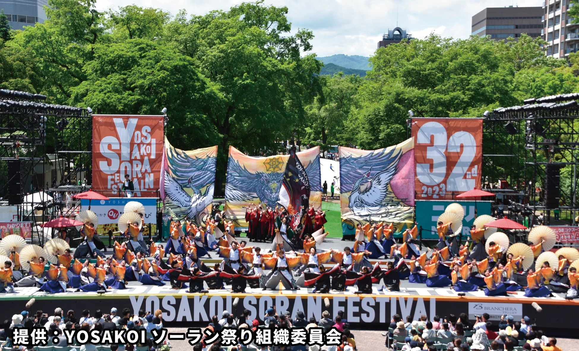 YOSAKOIソーラン祭りの屋外ステージで、カラフルな衣装をまとった大勢のダンサーが傘や扇子を手に踊っている様子